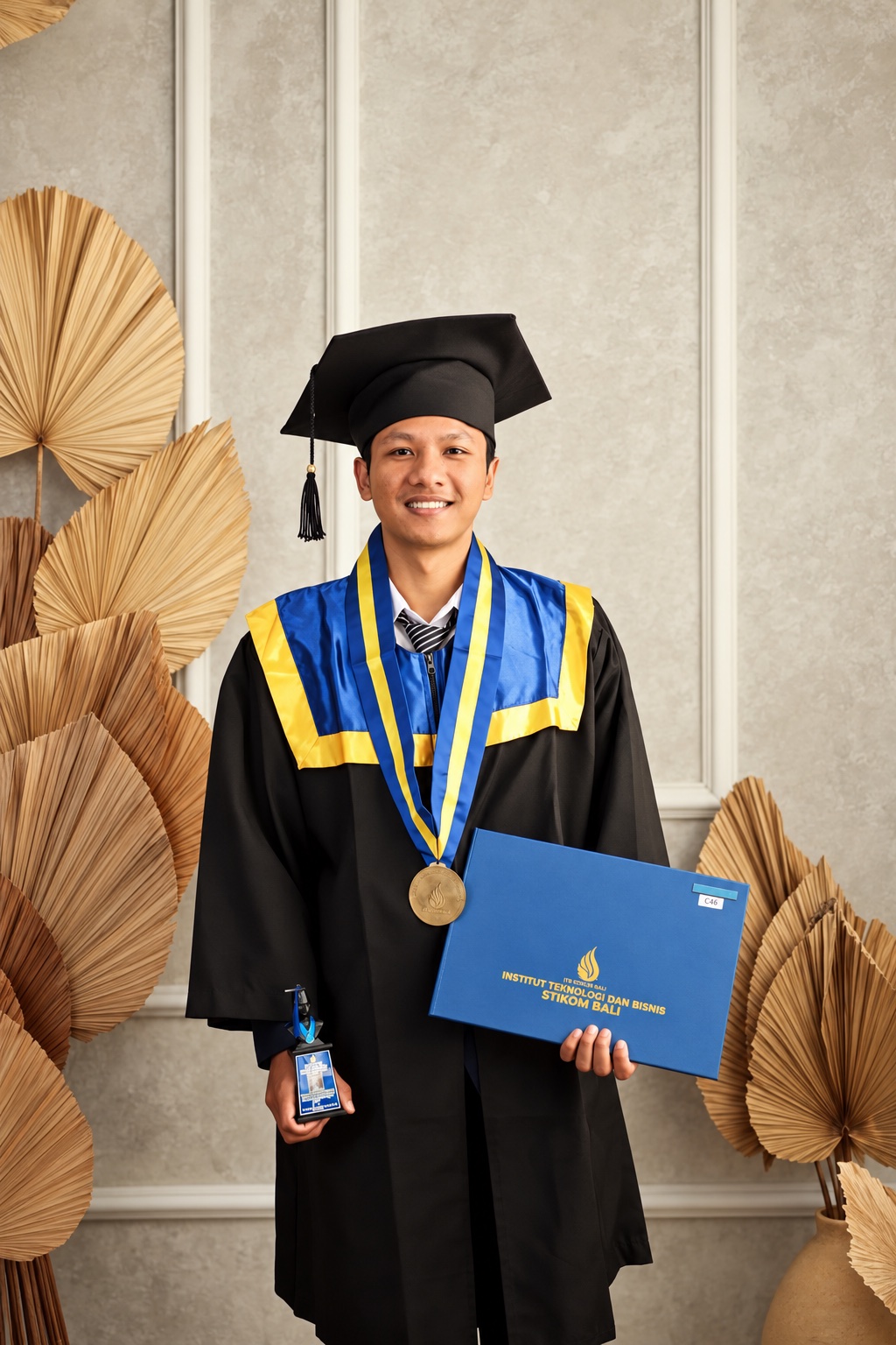 Wayan Widiarsana mengenakan toga wisuda dan memegang map Institut Teknologi dan Bisnis STIKOM Bali